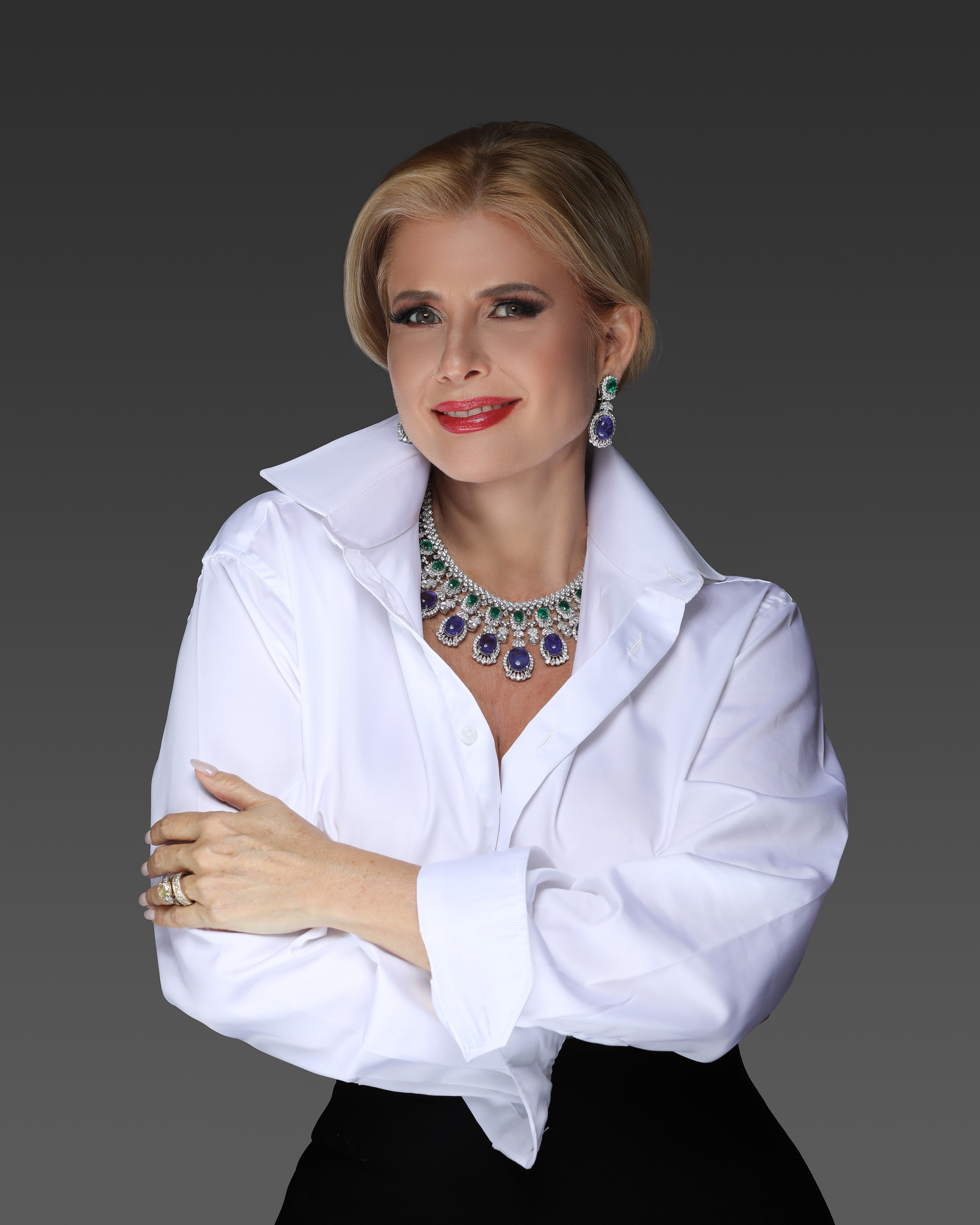 Cuban American actress Sissi Fleitas poses effortlessly with a blonde hair styled in an updo poses against a dark gradient background. She is wearing a crisp white button-down shirt with the collar popped and black pants. She accessorizes with a bold, statement necklace and matching earrings featuring large purple and green gemstones. She has red lipstick and subtle eye makeup, and her arms are crossed with a smile on her face.